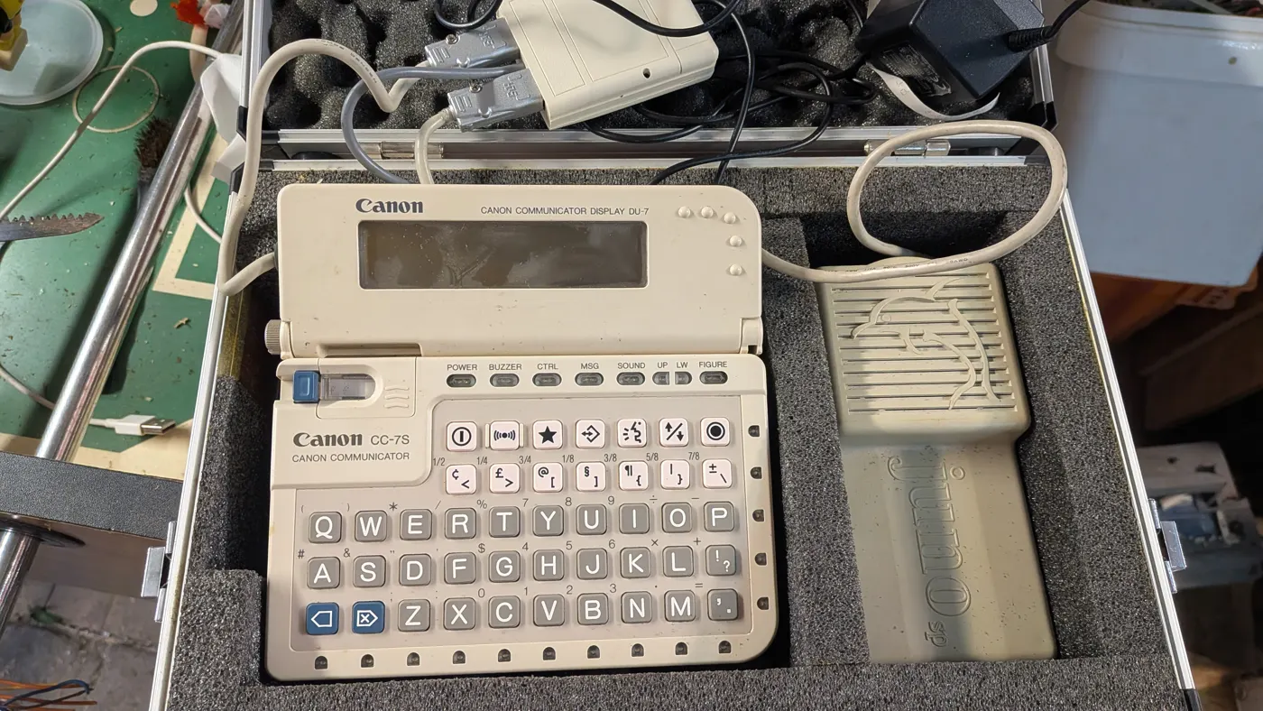 Geöffneter Koffer mit Canon Communicator CC-7S samt aufgeklapptem Display DU-7, daneben ein graues Zusatzgerät mit der Aufschrift Juno; im Deckel Kabel, Adapter und Netzteil